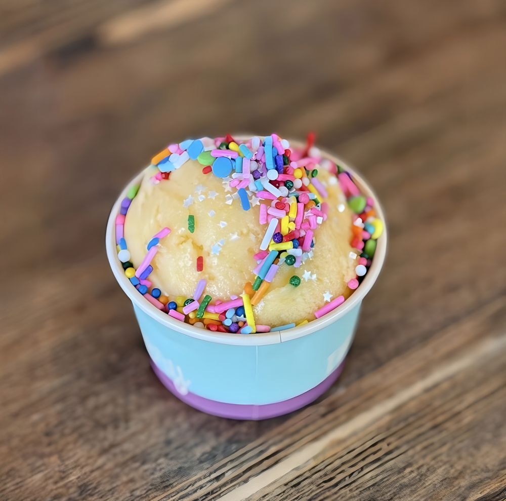 Cup of vanilla-colored dessert topped with colorful sprinkles on a wooden surface. 